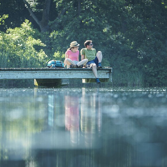 Ein Wanderpärchen sitzt auf dem Steg am Kratzmühlsee und lässt den Blick über den See schweifen. Hinter ihnen die Bäume und links am Steg ist ein Ruderboot befestigt.