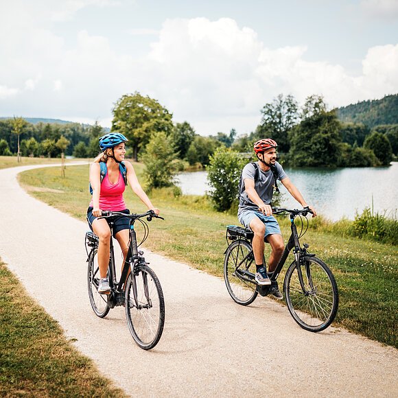 Eine Frau mit roten Shirt und blauem Fahrradhelm radelt neben einem Mann mit grauem Shirt und roten Helm. Rechts von ihnne liegt der Kratzmühlsee. Der Radweg ist von grünen Wiesen gesäumt. Der Himmel ist an diesem Tag etwas bewölkt.