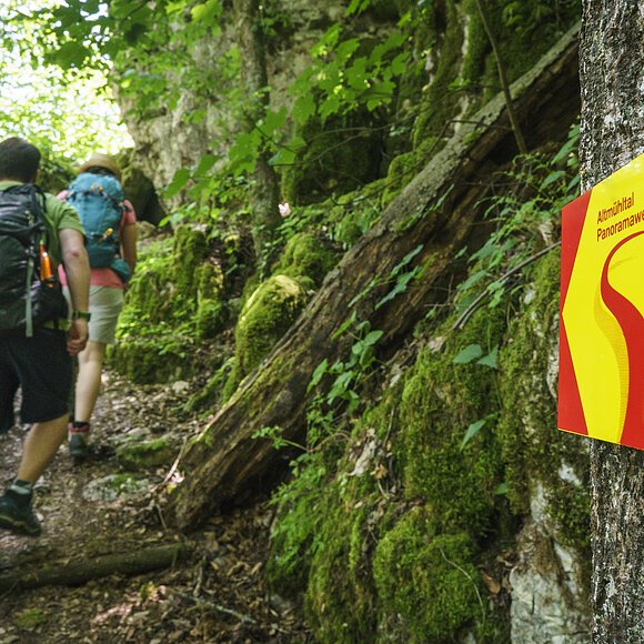 Im Vordergrund hängt ein Wanderschild des Altmühltal-Panoramaweges. Ein Wanderpärchen steigt die Treppen der Riedenburger Klamm hinauf.