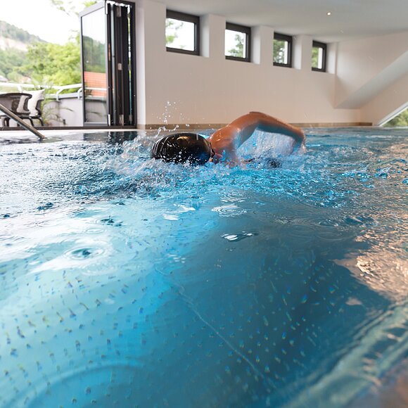 Ein Schwimmer zieht seine Bahnen im Becken. Im Hintergrund sind die Fenster mit dem Panoramablick zu sehen.