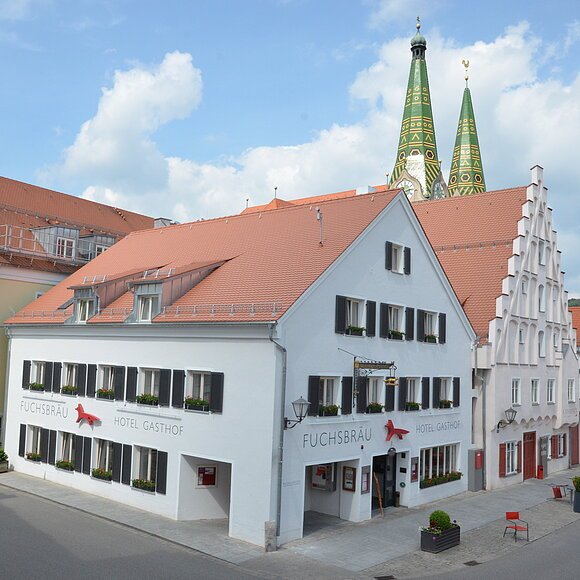 Hotel Fuchsbräu Beilngries Das Hotel an der Ecke ist weichgestrichen mit grünen Fensterläden. Im Hintergrund sind grüne Kirchtürme zu erkennen.