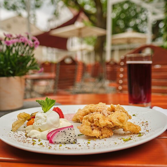 Brotzeit auf der Terrasse des Hotel Fuchsbräu Auf einem Holztisch auf der Terrasse ist eine köstlich angerichtete Portion Obatzda serviert. Im Hintergrund stehen links zwei kleine Blumentöpfe und rechts ein Getränk.