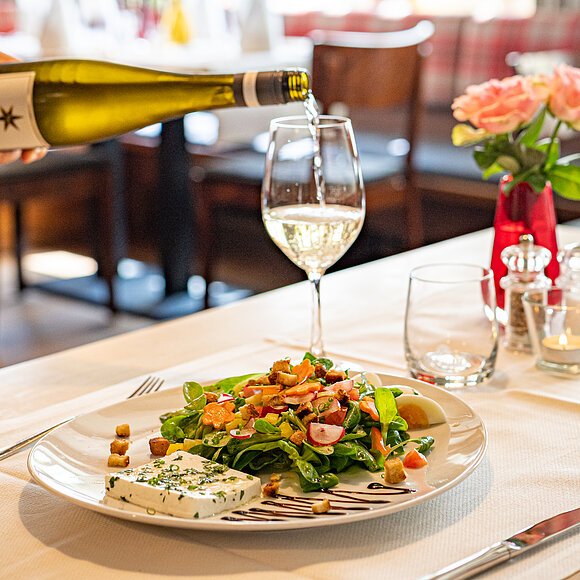Restaurant des Hotel Fuchsbräu Eine Hand gießt gerade Wein aus einer Flasche in ein Weinglas ein. Im Vordergrund steht ein Teller mit Salat und Käse.