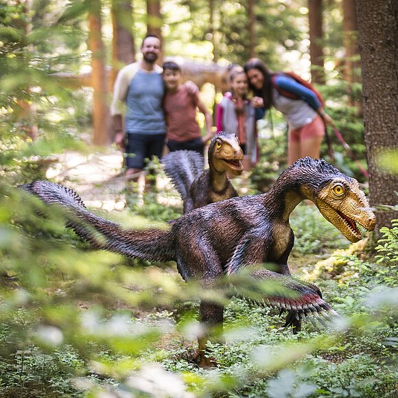 Zwei Dinosauriermodelle im Wald, unscharf im Hintergrund eine Gruppe von Menschen.