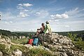 Zwei Wanderer sitzen auf einem Felsen und blicken auf bewaldete Hügel unter blauem Himmel mit Wolken.