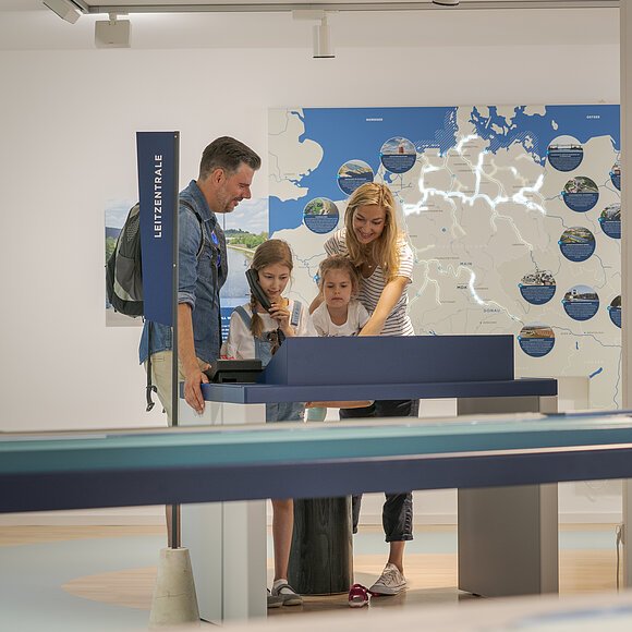 Familie mit zwei Kindern steht an interaktivem Ausstellungstisch vor beleuchteter Deutschlandkarte im Museum.