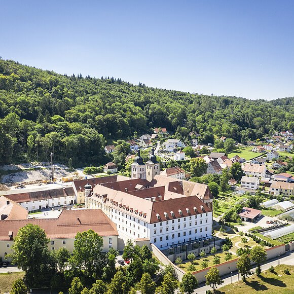 Luftaufnahme eines großen Gebäudekomplexes mit roten Dächern vor bewaldeten Hügeln und einem Dorf im Hintergrund.