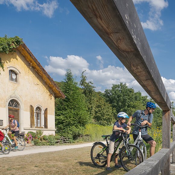 Familie mit vier Personen und Fahrrädern neben einem alten Haus und einem Fluss unter blauem Himmel.