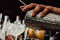 Zwei Gläser mit Eiswürfel stehen auf dem Tresen. Der Bartender gießt in das vordere den Cocktail in das Glas. Im Hintergrund sind die verschiedenen Flaschen zu erkennen.