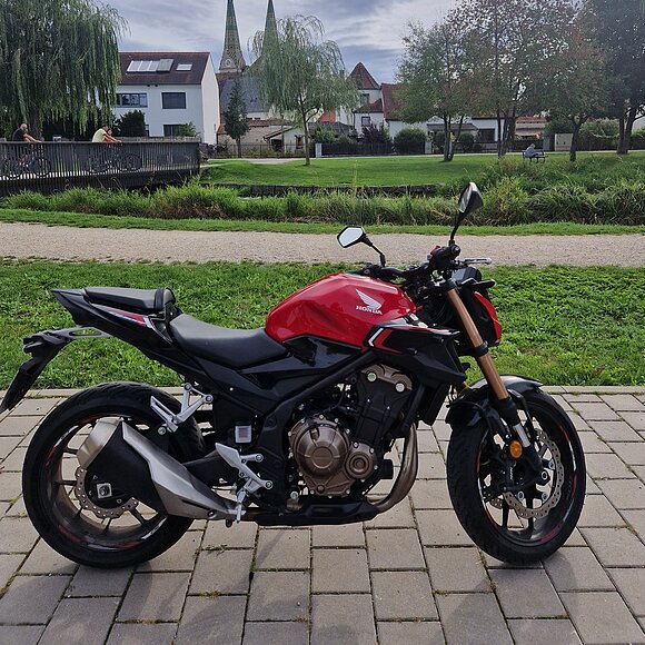 Motorrad der Marke Honda in rot und schwarz auf gepflastertem Weg vor Park mit Bäumen und Häusern im Hintergrund.