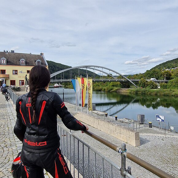 Person in schwarzer Motorradkleidung mit rotem Helm blickt auf Fluss, Brücke und Gebäude bei bewölktem Himmel.