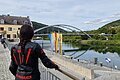 Motorradfahrerin macht Pause in Riedenburg Person in schwarzer Motorradkleidung mit rotem Helm blickt auf Fluss, Brücke und Gebäude bei bewölktem Himmel.