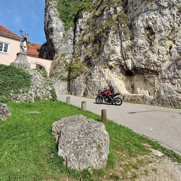 Motorradfahrer auf Straße vor steiler Felswand, links Statue und Gebäude mit rotem Dach.