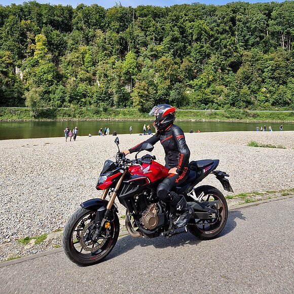 Person in schwarzer Motorradkleidung und Helm auf rotem Motorrad auf Weg am Flussufer mit Wald im Hintergrund