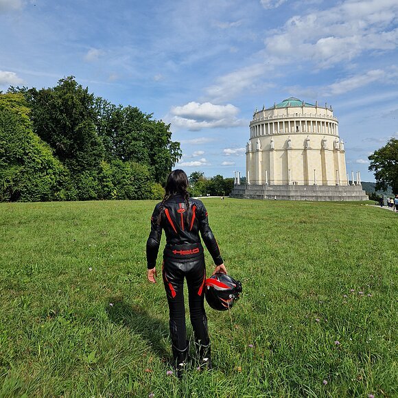 Person in schwarzer Motorradkleidung mit Helm in der Hand steht auf Wiese vor rundem Gebäude mit Säulen und Statuen.