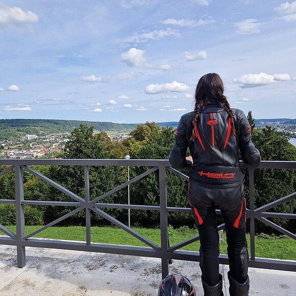 Motorradfahrerin bei Aussicht Befreiungshalle Kelheim Person in schwarzer Motorradkleidung mit roten Akzenten blickt von Balkon auf grüne Landschaft und Stadt, Helm liegt am Boden.