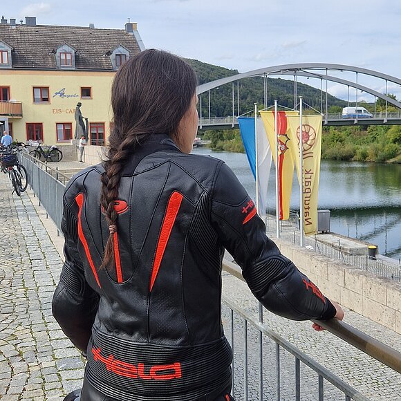 Motorradfahrerin in Riedenburg Person mit geflochtenem Haar und schwarzer Motorradjacke mit roten Akzenten lehnt an Geländer am Flussufer mit Brücke und Gebäuden im Hintergrund.