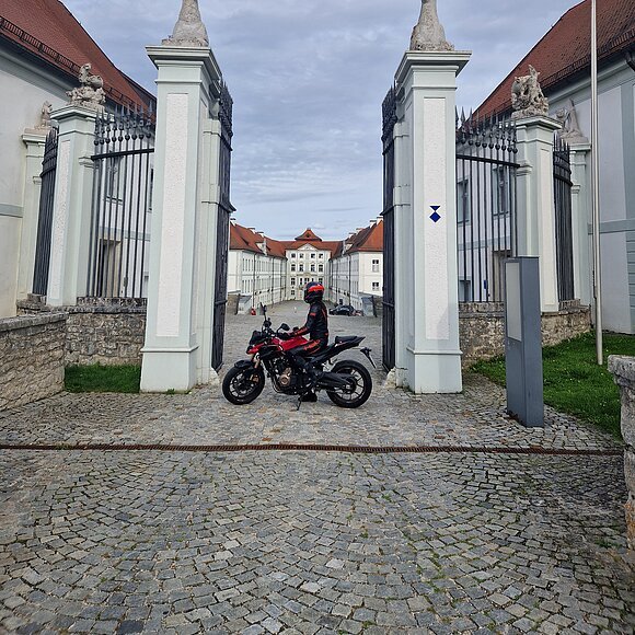 Motorradfahrerin vor dem Schloss Hirschberg Motorradfahrer in schwarzer und roter Schutzkleidung vor Tor mit Hirschstatuen und historischem Gebäude im Hintergrund.