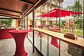 Veranstaltungsfläche im Freien im Hotel Fuchsbräu Überdachte Terrasse mit roten Stehtischen, Glaswand und Blick auf Garten mit rotem Sonnenschirm und Stühlen.