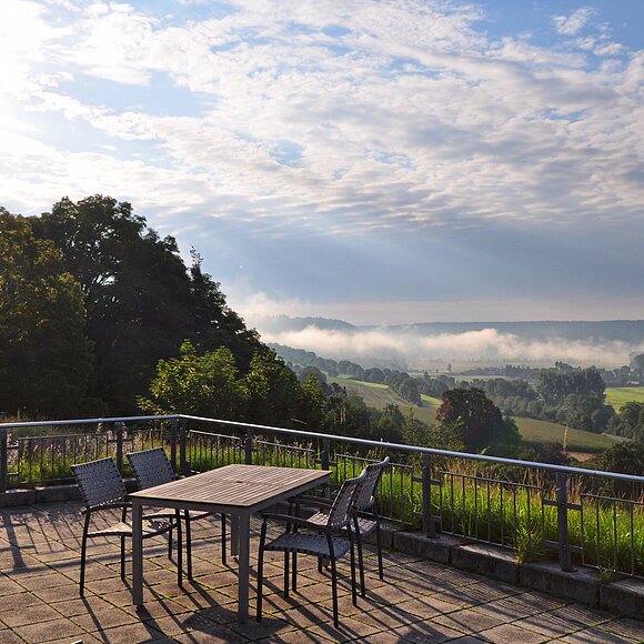 Terrasse vom ABG Tagungszentrum Terrasse mit Tisch und Stühlen, Blick auf neblige Landschaft mit Bäumen und Feldern bei bewölktem Himmel.