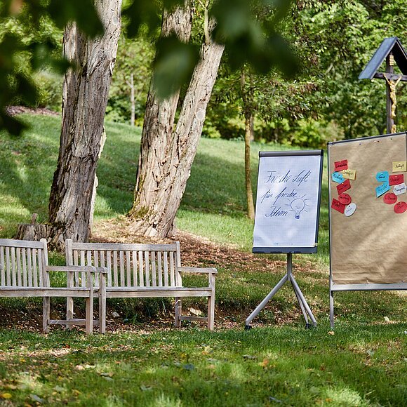 Tagungshaus AGB Beilngries draußen Zwei Holzbänke und zwei Flipcharts mit Notizen stehen auf einer Wiese unter Bäumen im Freien.