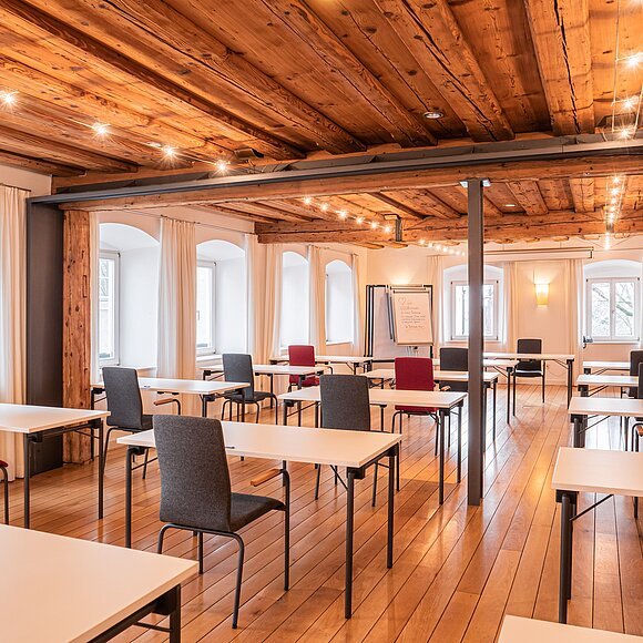 Historischer Saal im Hotel Fuchsbräu Seminarraum mit Holzboden, Holzbalkendecke, weißen Tischen und roten sowie grauen Stühlen, Fenster mit weißen Vorhängen.