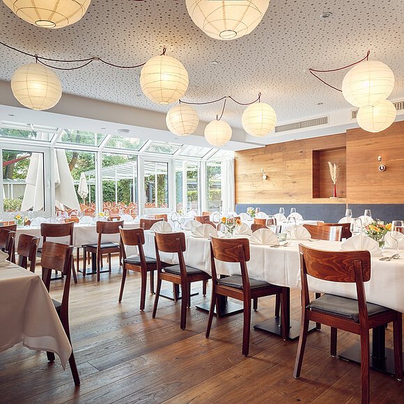 Veranstaltungssaal im Hotel Fuchsbräu Restaurant mit gedeckten Tischen, Holzstühlen, großen Fenstern und runden Papierlampen an der Decke.