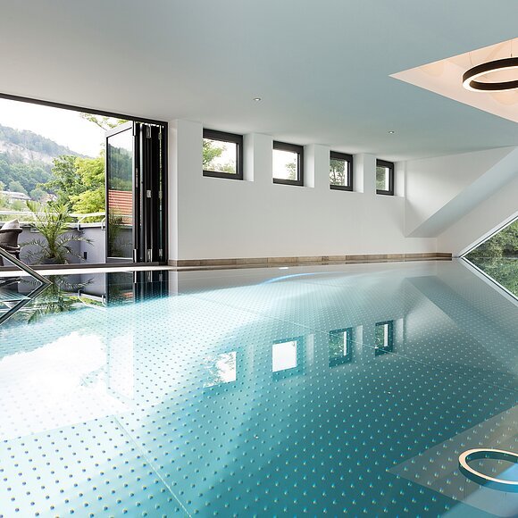 Innenraum eines modernen Hallenbads mit großen Fenstern und Blick auf eine Terrasse und bewaldete Hügel.