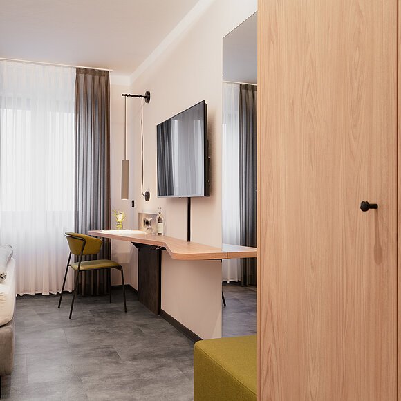 Hotelzimmer mit Bett, Schreibtisch, grünem Stuhl, Flachbildfernseher und Kleiderschrank aus Holz.