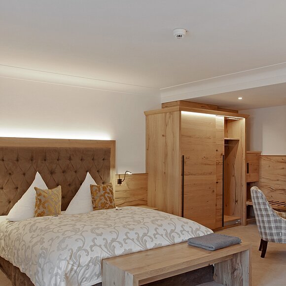 Hotelzimmer mit Doppelbett, Holzmöbeln, Sessel und Tisch in neutralen Farben und Beleuchtung.