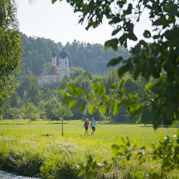 Ein Pärchen wandert unterhalb der Wallfahrtskirche Breitenbrunn entlang satter grüner Wiesen. Die Wallfahrtskirche ist von grünen Wäldern umgeben.