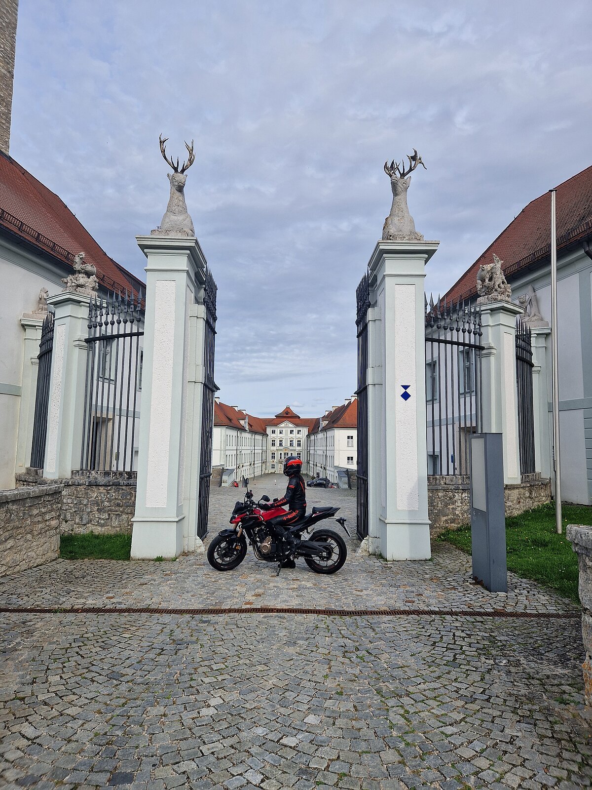 Motorradfahrer in schwarzer und roter Schutzkleidung vor Tor mit Hirschstatuen und historischem Gebäude im Hintergrund.