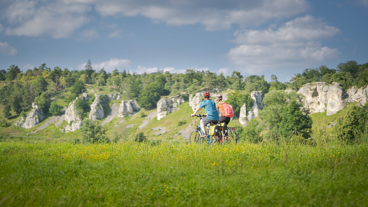 Zwei Personen fahren mit Fahrrädern auf einem grasbewachsenen Feld vor einer Felslandschaft.