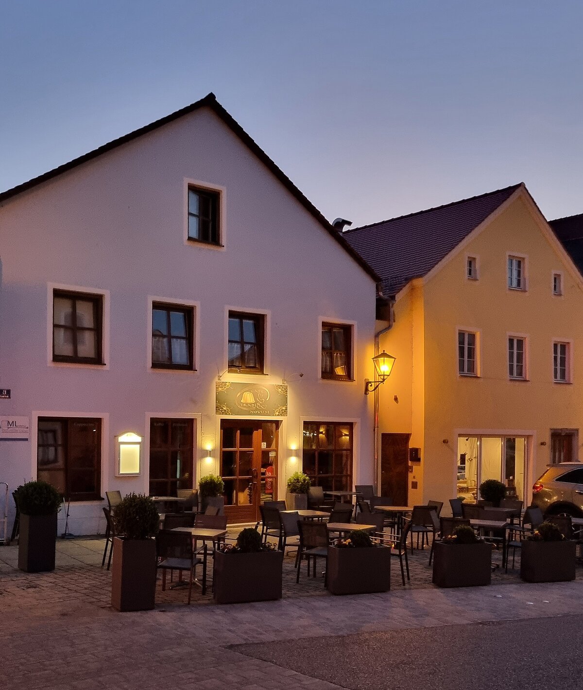 Das Bild zeigt die Außenterrasse des Café Tradinovum bei Dämmerung. Das Café hat eine weiße Fassade und wird von warmem Licht der Straßenlaternen beleuchtet. Vor dem Café sind Tische mit dunklen Stühlen und Pflanzgefäßen auf einem grauen Steinpflaster angeordnet, was eine einladende Atmosphäre schafft.