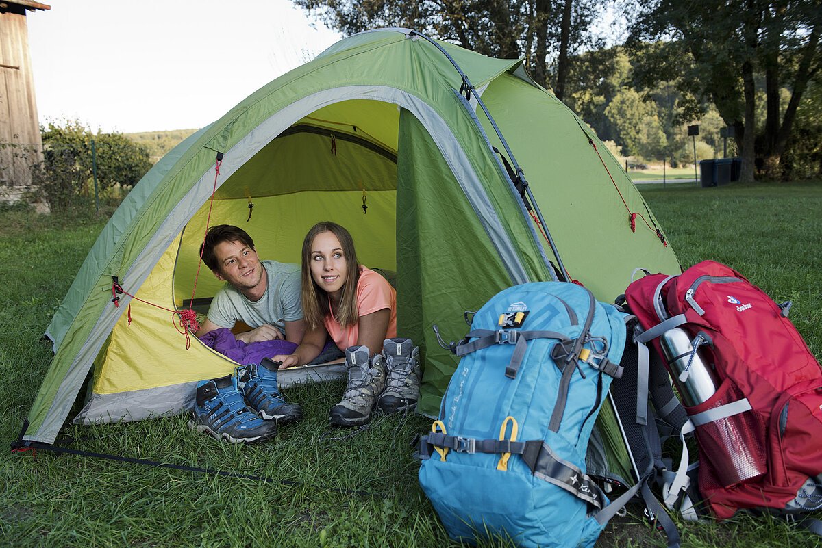 Zwei Personen liegen in einem grünen Zelt, davor Wanderschuhe und zwei große Rucksäcke auf Gras.