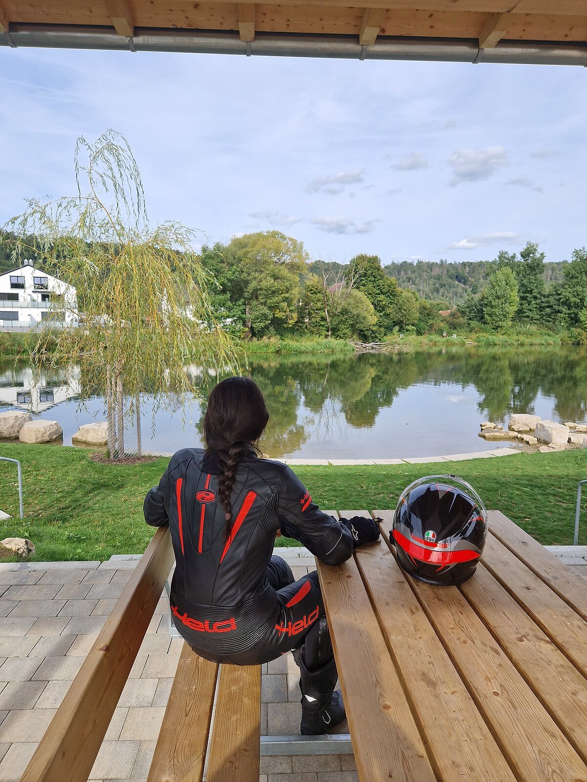 Person in schwarzer Motorradkleidung sitzt auf Holzbänken unter einem Dach und blickt auf einen See mit Bäumen im Hintergrund.