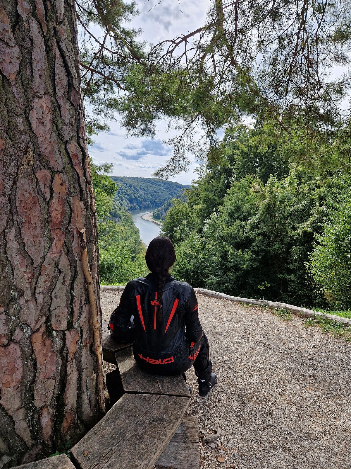 Motorradfahrerin Person mit schwarzer Motorradjacke sitzt auf Holzbänken und blickt auf Fluss und bewaldete Hügel.