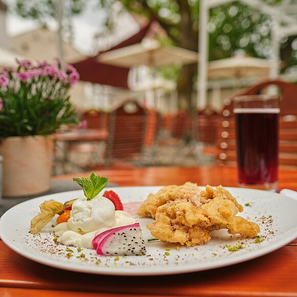 Auf einem Holztisch auf der Terrasse ist eine köstlich angerichtete Portion Obatzda serviert. Im Hintergrund stehen links zwei kleine Blumentöpfe und rechts ein Getränk.