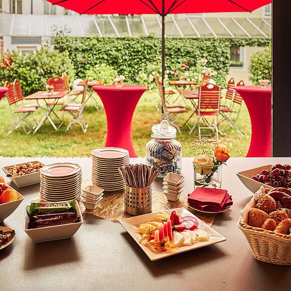 Buffet mit Obst, Gebäck, Nüssen und Tellern auf Tisch vor Garten mit roten Stehtischen und Stühlen
