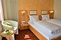 Hotelzimmer mit zwei Einzelbetten, Nachttisch, Sessel, kleinem Tisch und Obstschale bei Tageslicht.
