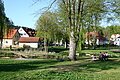 Grüne Parkwiese mit Bäumen, Bach, Fahrrad und Menschen auf Bank vor Häusern unter blauem Himmel.